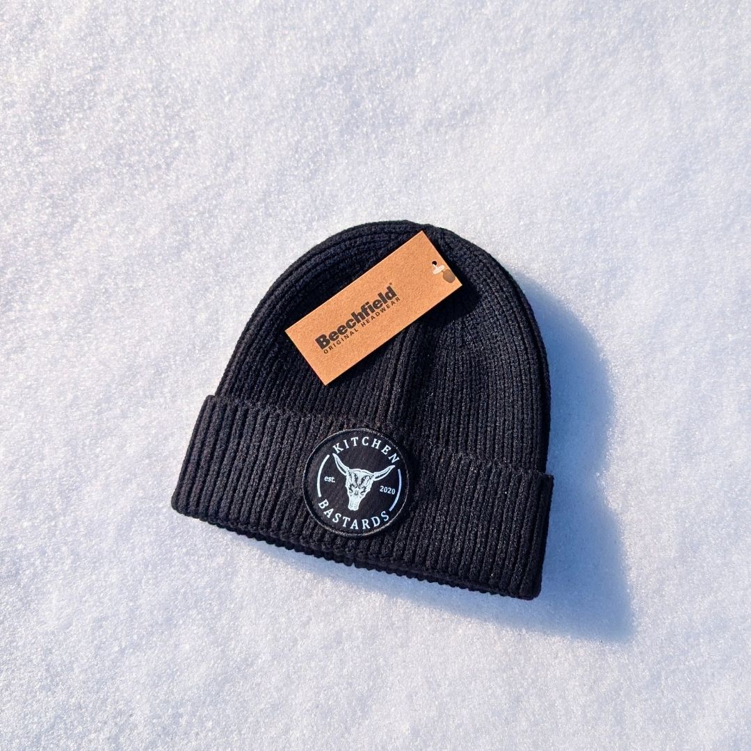 Schwarze Beanie mit Kitchen Bastards Logo auf weißem Untergrund im Schnee